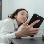 Caucasian woman relaxing in bed, holding mobile phone in morning light.